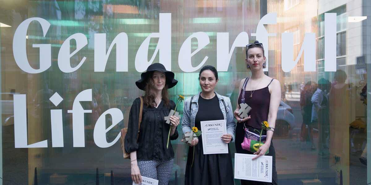 Drei Personen stehen vor einem halbtransparenten Schaufenster mit dem Schriftzug "Genderful Life". In den Händen halten sie je eine Urkunde, eine Sonnenblume und ein graues Ungleichzeichen.