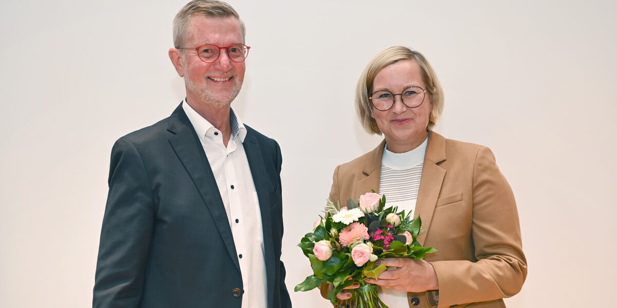 Eine männliche gelesene Person steht neben einer weiblich gelesener Person, die einen Blumenstrauß in der Hand hält.