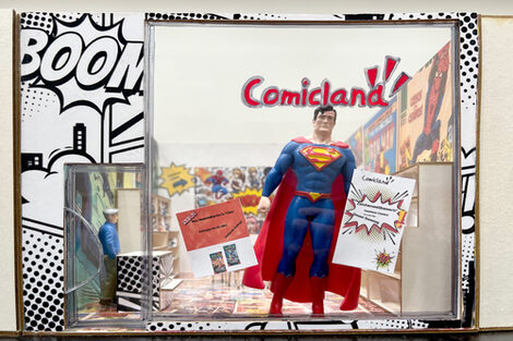 A Superman figure stands in the window of a miniature model of a comic store.