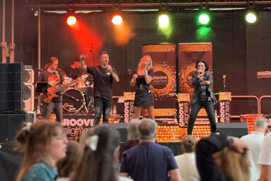 Frontalblick über die Köpfe einiger Menschen hinweg auf eine Bühne, auf der ein Bassist, ein Schlagzeuger und drei Sänger*innen in Aktion zu sehen sind.