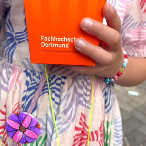 Eine Kinderhand hält eine orangefarbene Popcornschachtel, das Kind trägt ein buntes Armband und einen Buttom am Kleid.