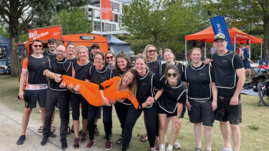 15 people in black jerseys carry another person in orange on their hands, stand next to each other and look into the camera. In the background, a car and two tents, both orange, stand on a meadow.