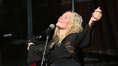 Eine Person mit langen blonden Haaren und ausgestreckten Armen steht mit geschlossenen Augen und nach innen gerichtetem Lächeln vor einem Mikrofon.