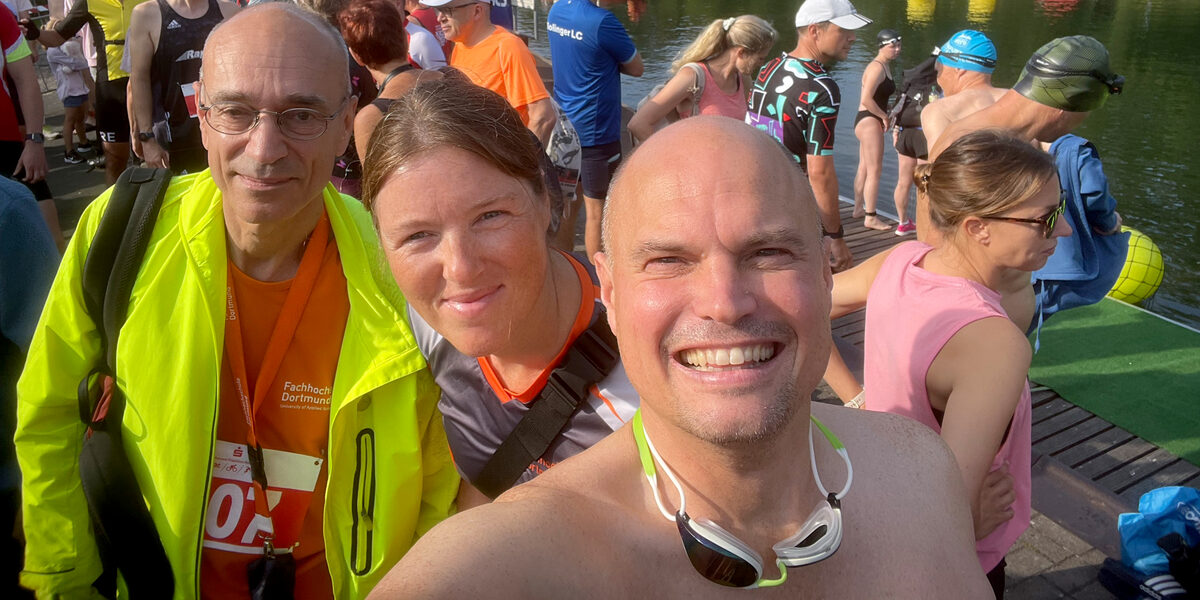 Selfie of three people under an open summer sky with many other people in the background on the bank of a watercourse.