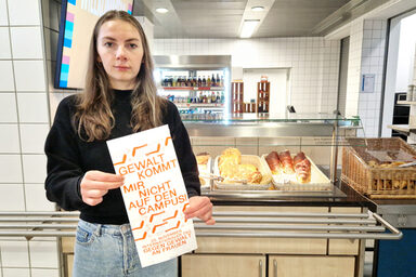 Eine Person steht in der Mensa vor einer Theke mit Backwaren und hält eine Tüte vor sich mit der Aufschrift: "Gewalt kommt mir nicht auf den Campus!".