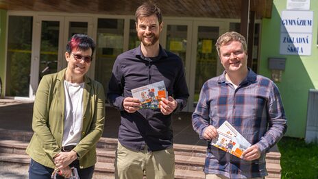 Drei Personen stehen vor einer Schule und halten Flyer der FH Dortmund in den Händen.