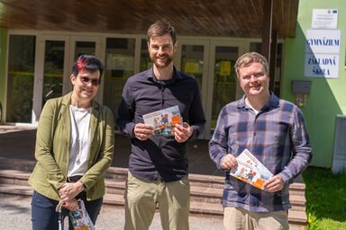 Drei Personen stehen vor einer Schule und halten Flyer der FH Dortmund in den Händen.