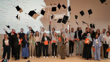 Eine Gruppe von Personen steht auf einer Bühne und wirft schwarze Doktorhüte als Symbol für den Abschluss in die Luft. Die Personen tragen unterschiedliche Kleidung, darunter Anzüge, Kleider und legere Outfits. Einige halten orangefarbene Dokumentenmappen in der Hand. Im Hintergrund ist eine weiße Wand mit einem abstrakten Muster und Texten zu sehen. Vor der Bühne sind Zuschauer mit Kameras zu erkennen, die Fotos machen.