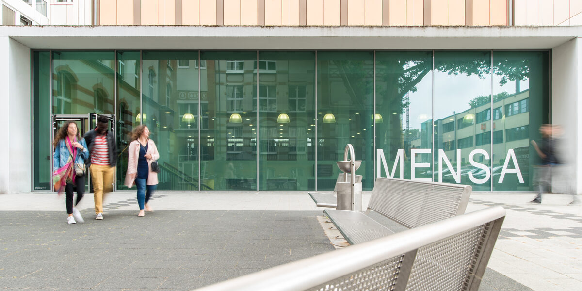 Foto des Mensagebäudes, im Vordergrund Bänke. Vor der Mensa verschwommen vier Personen.__The canteen building, benches in the foreground. Four people blurred in front of the canteen.