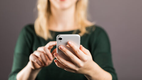 Junge Frau - Kopf oben im Anschnitt - hält vor sich ein Smartphone in der linken Hand, mit dem Zeigefinger der rechten Hand berührt sie fast das Display.