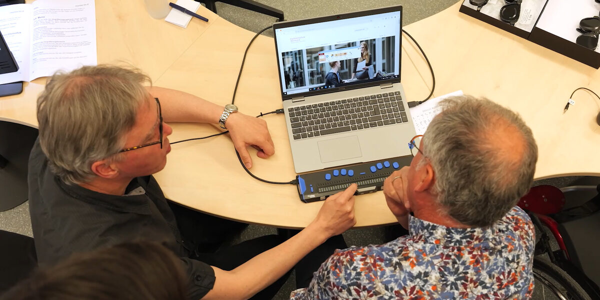 Zwei Personen sitzen an einem Tisch. Und schauen auf einen Laptop. Eine Person zeigt der anderen eine Vorrichtung, mit denen blinde Menschen die Inhalte des Bildschirms erfassen können.