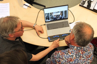 Zwei Personen sitzen an einem Tisch. Und schauen auf einen Laptop. Eine Person zeigt der anderen eine Vorrichtung, mit denen blinde Menschen die Inhalte des Bildschirms erfassen können.