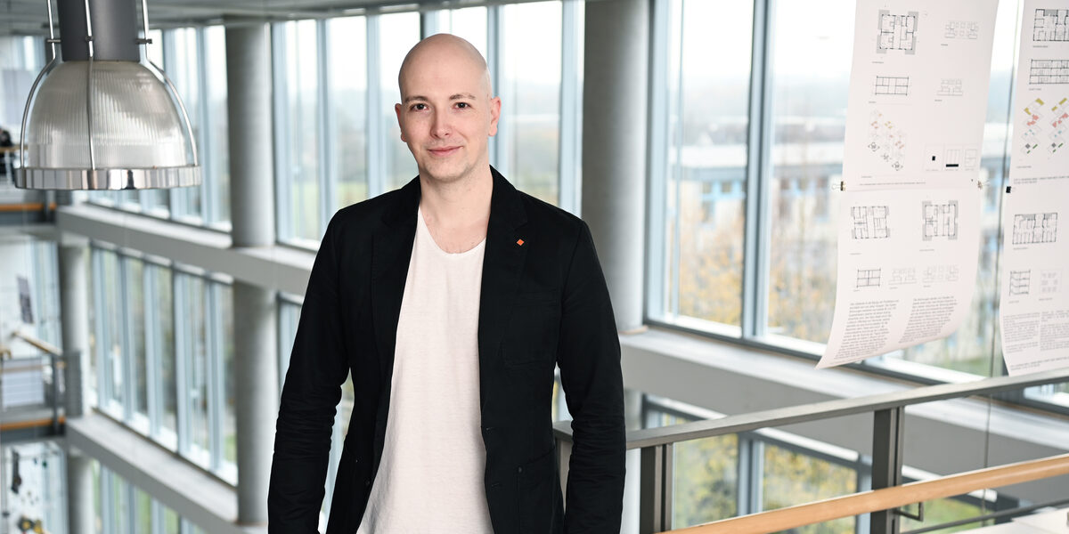 Ein Mensch im schwarzen Sakko steht in einem Gebäude an einem Geländer vor einer Fensterfront.