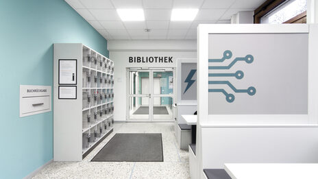 Foto des Flurs vor dem Eingang zur Bibliothek. Dort befinden sich Schließfächer, eine Rückgabebox und Gruppenarbeitsplätze__Photo of the hallway in front of the library entrance. It features lockers, a book return box and desks for group study