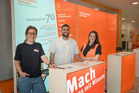 Drei Personen stehen vor einer orangefarbenen Wand auf der „Mach was mit Wissen“ steht. Sie lächeln.