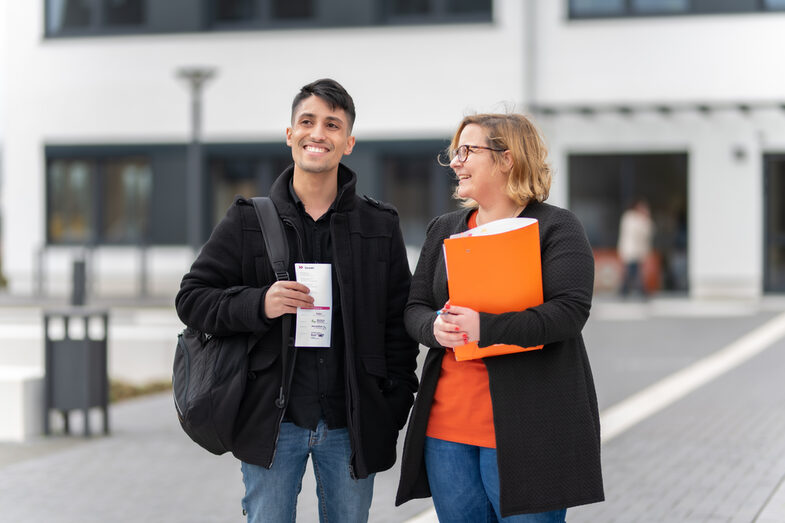 Photo of an employee and a student standing in front of a campus building. She is holding an orange folder under her arm, he has a flyer in his hand. They are both laughing.