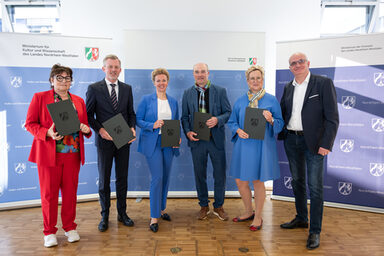 Sechs Personen stehen nebeneinander, fünf von ihnen halten eine Mappe vor sich.