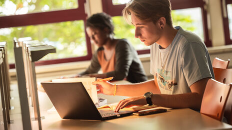 Foto von zwei Studierenden, die an einem Lernplatz in der Bibliothek nebeneinander sitzen und lernen.