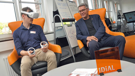 Two people are sitting on two armchairs next to each other. The UAS employee on the left has put on VR glasses and is holding two control elements, while the Lord Mayor looks on.