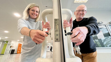 Two people are each standing on one side of a door in a building, holding the door handle with one hand and a key in the lock of the door with the other. Both are smiling and looking at the camera.