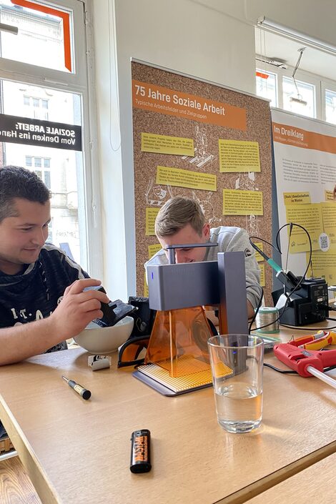 Zwei Männer werkeln an einem kleinen Brunnen. Vor ihnen liegen Werkzeuge auf einem Tisch.