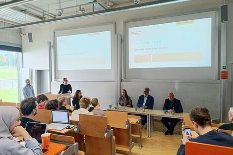 Zu sehen sind die drei Gäste in der Vorlesung Politisches Denken - Politisches Handeln , die an einem Tisch sitzen. Daneben stehen die Moderator*innen