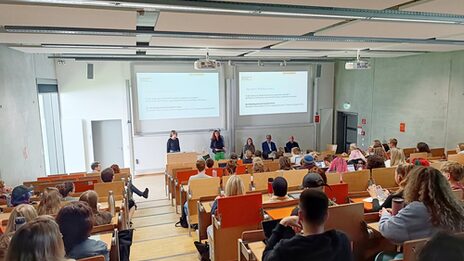Vorlesung im Hörsaal U33, in der der Saal voll ist und im Vordergrund die Professorin Ute Fischer Gäste vorstellt