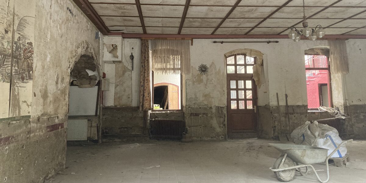 In einer leeren Wohnung steht eine Schubkarre, die verdreckten Wände und zerstörten Fenster lassen den Raum wie eine Baustelle aussehen.