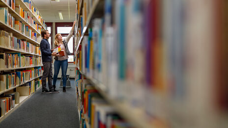 Foto einer Studentin und eines Studenten, die in einer Regalreihe in der Bibliothek Emil-Figge-Straße stehen und Bücher aus dem Regal holen.__A female and male student stand in a row of shelves in the Emil-Figge-Strasse library, fetching books from the shelf.