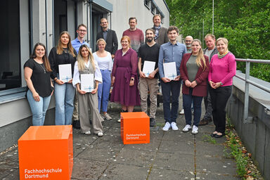 Eine Gruppe von Menschen steht um orangefarbene Würfel. Einige von ihnen halten Urkunden.