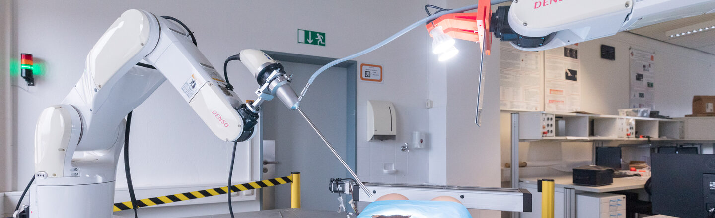 Photo of a surgical set-up with body and two surgical robots in action. The one on the right has an endoscope, the one on the left has light bulbs attached. Behind it you can see desks in the laboratory.
