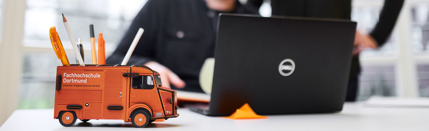 Foto einer Stiftebox in Form des Kastenwagens der Fachhochschule auf einem Schreibtisch. Unscharf dahinter sitzt ein Mann am Laptop und eine Frau steht hinter ihm, beide schauen auf den Bildschirm.