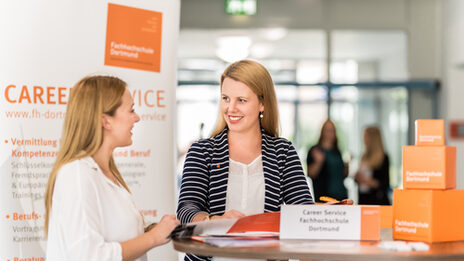 Foto von einer Mitarbeiterin des Career Services der Fachhochschule Dortmund, die an einem Stehtisch im Gespräch mit einer jungen Frau steht. __ <br>Employee of the Career Services of the University of Applied Sciences Dortmund stands at a high table talking to a young woman.