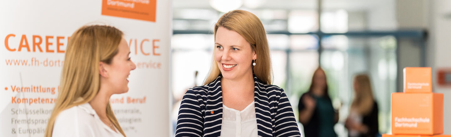 Foto von einer Mitarbeiterin des Career Services der Fachhochschule Dortmund, die an einem Stehtisch im Gespräch mit einer jungen Frau steht. __ <br>Employee of the Career Services of the University of Applied Sciences Dortmund stands at a high table talking to a young woman.