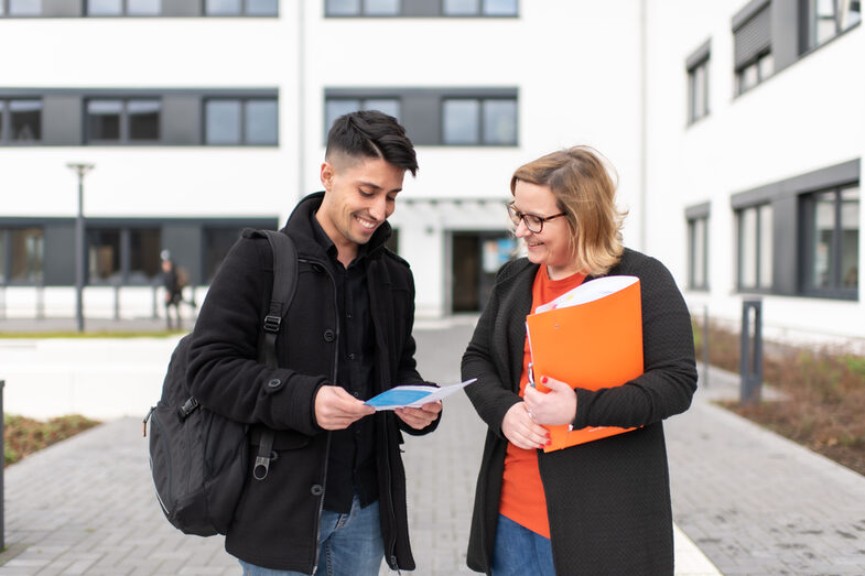 Foto einer Mitarbeiterin und eines Schülers, dir vor einem Campusgebäude stehen. Sie hält eine orangene Mappe unterm Arm, er hat einen Flyer in der Hand. Beide schauen auf den Flyer und lachen.