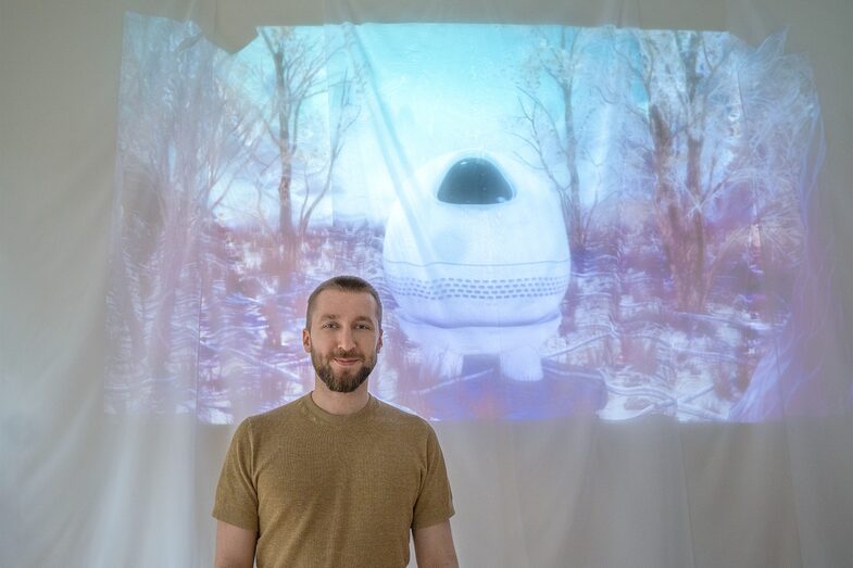 Lennart Gruensel stands smiling next to the projection of Argos, an artificial intelligence.