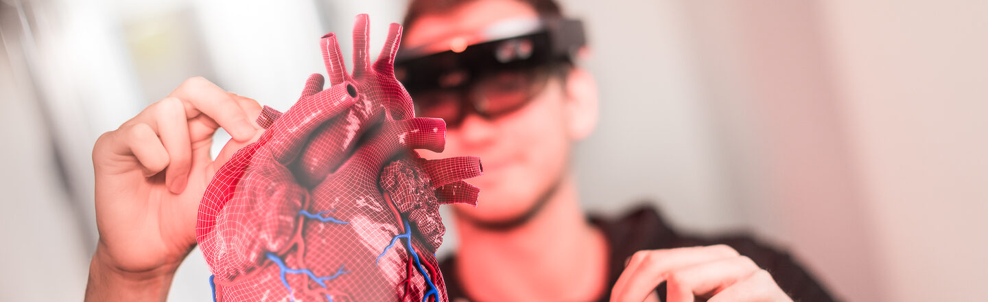 Photo of a student wearing VR goggles, holding his hands up in the air for control. In front of him you can see what he sees through the glasses: a 3D heart hollogram. __ Student wears VR glasses and holds hands up to control. In front of him you can see what he sees through the glasses: a 3D heart hollogram.