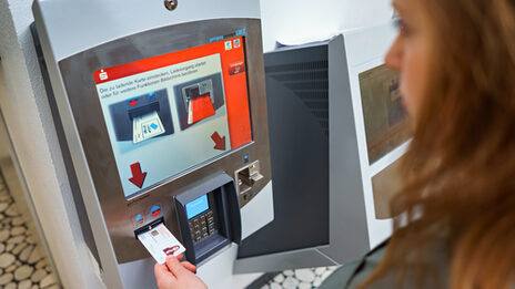 Foto einer Studentin, die ihren Studierendenausweis zum Aufladen in den Automaten der Sparkasse steckt.__A student puts her student ID card into the Sparkasse's machine to top up.