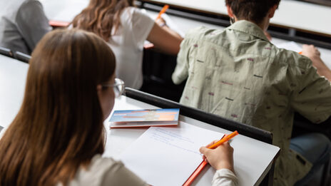 Photo of a young woman taking notes in the lecture hall. Three other people are seated in front of her. __ Young woman takes notes in the lecture hall, three other people are seated in front of her.