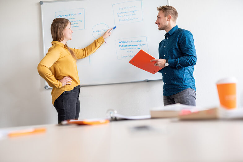 Two students are standing in front of a board on which a graphic of the "marketing mix" is drawn. The student on the left of the picture points to a "box" and looks at her fellow student.