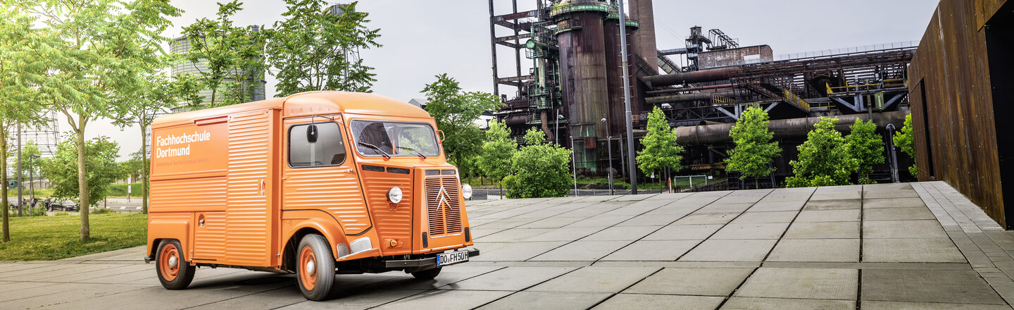 Photo of orange vintage car with FH logo in front of an industrial monument ___Orange vintage car with FH logo in front of an industrial monument.