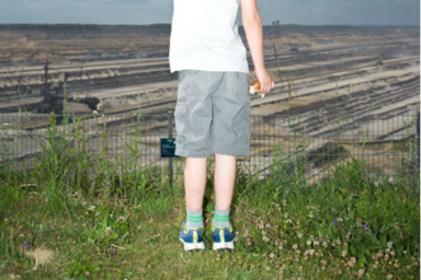 Ein junger Mensch, von hinten fotografiert, steht in T-Shirt und kurzer Hose auf einem Stück Wiese direkt vor einem Zaun. Hinter dem Zaun erstreckt sich ein Abbaugebiet bis zum Horizont.