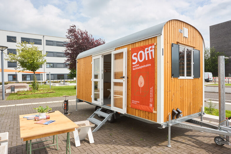 You can see a construction trailer with a poster saying "Soffi mobile sustainability office"
In front of the trailer you can see a table with a few things on it.