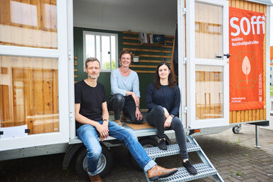 Man sieht 3 Personen, die in einem Bauwagen sitzen, welchen man über eine kleine Treppe betreten. An dem Bauwagen ist ein großes Plakat befestigt auf dem „Soffi mobiles Nachhaltigkeitsbüro“ steht.