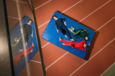 The photo shows three people in tracksuits lying on a sports mat.