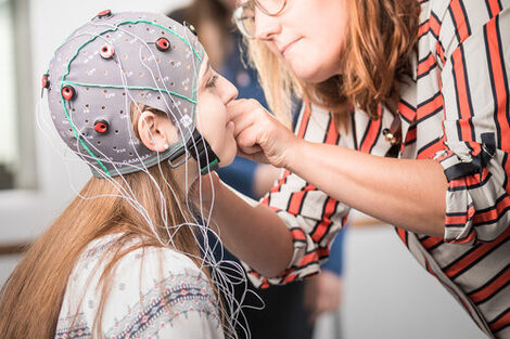 Foto einer Studentin, der eine EEG-Maske von einer Frau in einer orange gestreiften Bluse angezogen wird.