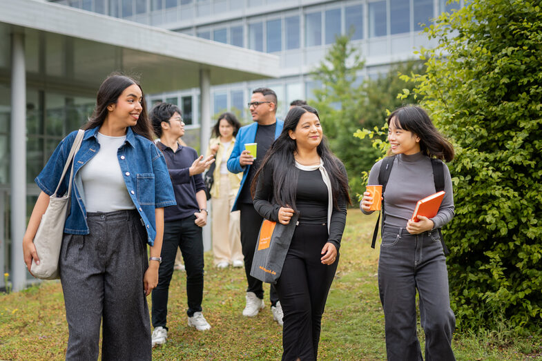 Gruppe internationaler Studierender läuft mit Taschen und Getränkebechern vor Hochschulgebäuden gemeinsam über eine Wiese des Campus Emil-Figge-Straße.