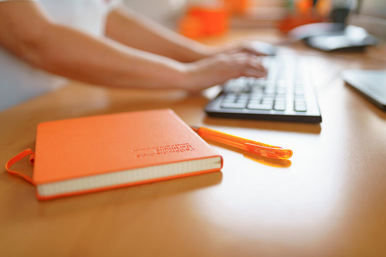 Foto von einem FH-Notizbuch und FH-Kugelschreiber, die auf einem Schreibtisch liegen. Im Hintergrund tippt eine Frau auf der Tastatur. __ FH notebook and FH ballpoint pen are on the desk, a woman is typing on the keyboard in the background.